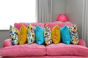 Multi colored cushions lined up in a row on a bright pink couch with light grey walls and curtains plus pink lampshade behind.	