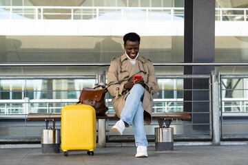 Smiling Afro-American traveler man with yellow suitcase sitting on bench in airport terminal or...