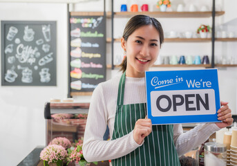 Beautiful asian young barista woman in apron holding and showing open sign board in cafe. Happy and smile barista girl welcome to front counter bar in coffee shop. Business owner startup concept.