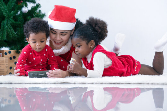 Happy Family, Mom With Her Dark Skinned Daughter And Son Lying On Floor And Looking At Something At Smart Phone Near Christmas Tree Indoors, Merry Christmas