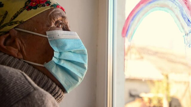 An old grandmother on self-isolation during quarantine. A masked grandmother looks out the window during self-isolation. Grandma looks out the window through a rainbow pattern during the Covid-19