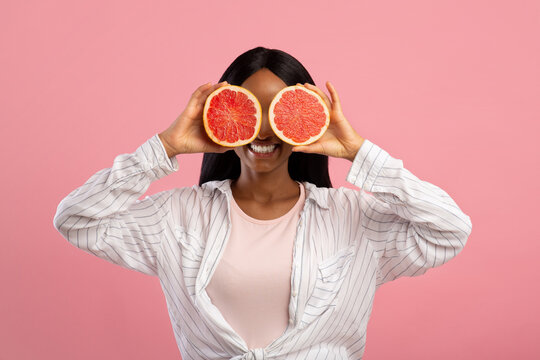 Vitamins for beauty and skin care. Happy African American woman covering eyes with grapefruit halves on pink background
