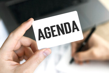 Text AGENDA on card in hand on the background of laptop and documents.