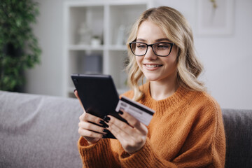 Happy young woman in eyeglasses using digital tablet and credit card for online shopping while staying at home. Concept of modern technology.