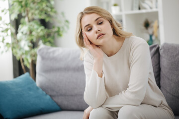 Obraz premium Tired young woman with hairstyle sitting on grey sofa with closed eyes. Bored lady in white clothing sitting at home during leisure time.