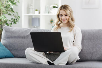 Beautiful young blonde sitting with wireless laptop on couch and surfing internet. Pretty woman...