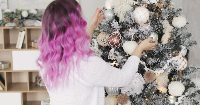 New Year, Christmas, Xmas, holiday and celebration concept. Young woman with pink hairs is decorating Christmas tree with balls, garlands and pompons. She is standing back to camera.