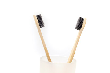 Two bamboo wooden eco toothbrushes with charcoal coating in glass cup on white background.