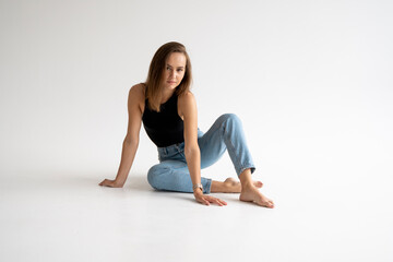 Fototapeta na wymiar Portrait of happy young pensive woman posing in black underwear and blue jeans without a shoes, sitting on a white floor in white studio. Model tests of pretty girl in basic clothes on cyclorama.