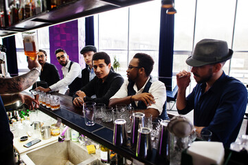 Group of handsome retro well-dressed man gangsters spend time at club. Drinking whiskey at bar counter. Multiethnic male bachelor mafia party in restaurant.