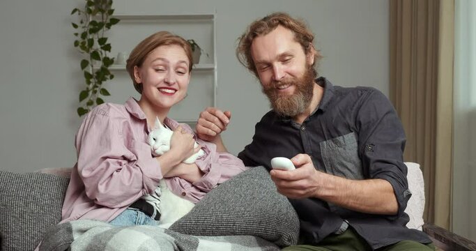 Caucasian couple blonde woman and bearded man sitting with white fluffy cat on couch in living room watching TV movies relaxing on sofa husband switches channel while his wife strokes his beloved pet