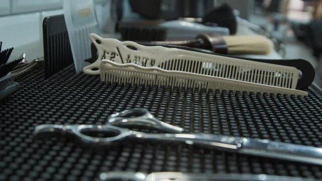 The hand of the barber takes (chrome) scissors from the (black rubber) mat on which the (black and white) combs and the straight razor lie