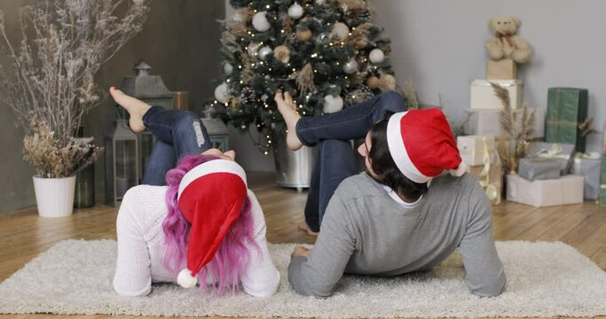 New Year, Christmas, Xmas, holiday and celebration concept. Happy young couple in Santa's red hats is lying together near Christmas tree, back view. Family lovely moments in magic time.
