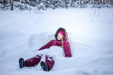 Fototapeta na wymiar A girl in a red jumpsuit sits on the snow and looks with hope and surprise into the distance against the background. The concept of the winter holidays.
