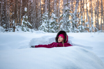 A girl in a red jumpsuit sits on the snow and looks with hope and surprise into the distance against the background of the forest and the sunset. The concept of the winter holidays.