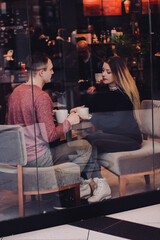 Fototapeta na wymiar Cute brunette loving couple in cozy warm sweaters on a date at the cafe looking at the phone, using it. cold autumn, winter or spring day, european city. indoors