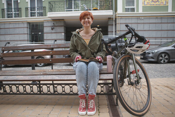 Attractive happy young woman laughing joyfully, sitting on a bench near her bicycle, reading a book, copy space