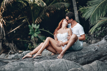 Happy young romantic couple in a white summer clothes love posing at the camera  standing on an old tree under palm trees on the beach. Red haired girl and handsome man in a summer vacation. Phuket. T