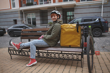 Plakat Full length shot of a happy young female delivery courier resting on a bench in the city after riding bicycle with her delivery backpack