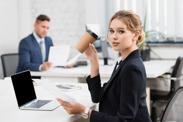 Businesswoman with paper cup looking at camera, while holding paper sheet at workplace on blurred 