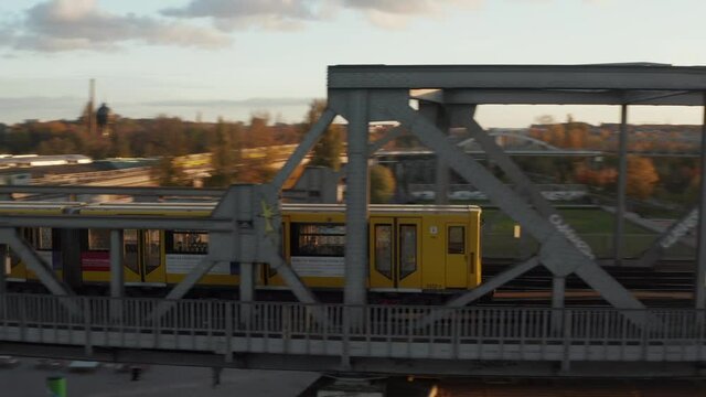 Yellow Subway Train passing bridge through urban environment in Berlin, Germany in beautiful golden hour Sunset light, Aerial tracking follow shot