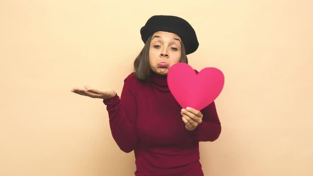 Young latin woman with boina holding heart shrugs shoulders and open eyes confused