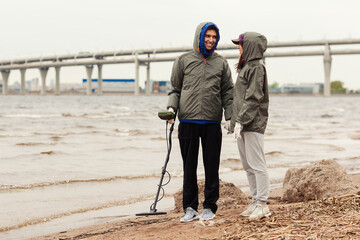 Happy loving young couple in hoods standing near coastline holding hands and using metal detector...