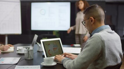 Medium shot of team of colleagues sitting at table together working on laptops and listening to young mixed race woman making report on new business project next to projector in blurred background