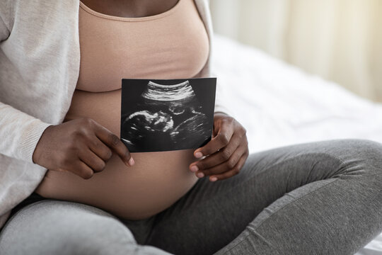Pregnant black woman sitting on bed holding ultrasound image of baby, closeup