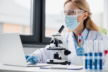 scientist in medical mask and goggles holding test tube with covid lettering and using laptop, stock image