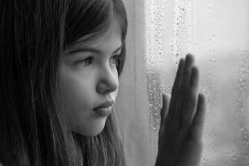 little, young sad girl at the window. Keeps his hand on the glass, lonely child depressed
