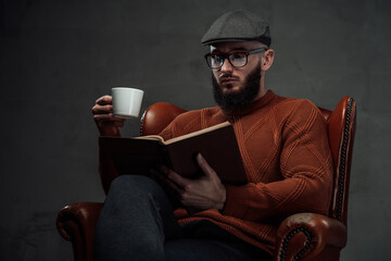 Sitting on leather armchair in dark background clever and manly guy with beard and glasses weared in sweater with cap. - obrazy, fototapety, plakaty
