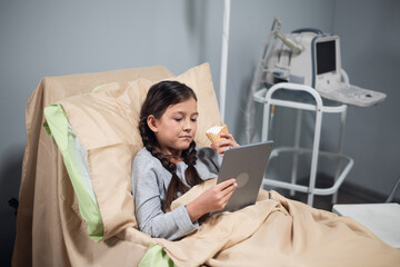 Positive little girl using an app on her digital device, while staying in a hospital bed.