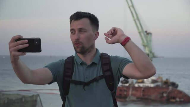 Young traveler blogger takes selfie on phone, makes video broadcast for subscribers on Sea coast about sunken tanker Delf, its rescue using floating crane. Shooting vlog, streaming online podcast