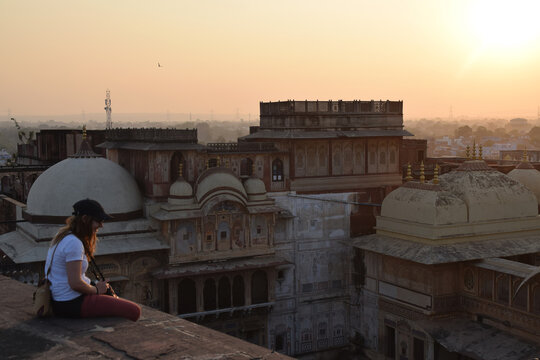 Young woman and tourist, sitting on the rooftop and is watching the sunset over the city skyline with a camera around her neck. Photo taken in India.