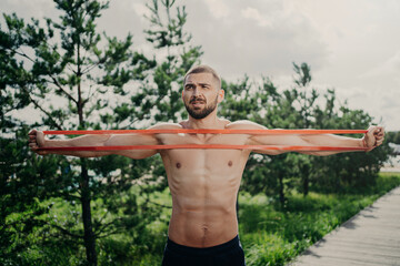 Horizontal shot of sporty man with bare chest exercises with elastic expander, gains for strong...