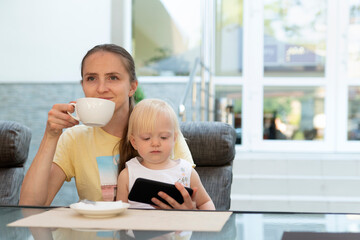 Happy young mom drinks coffee and holds child in her arms. Kid watching cartoons on phone while mom is relax