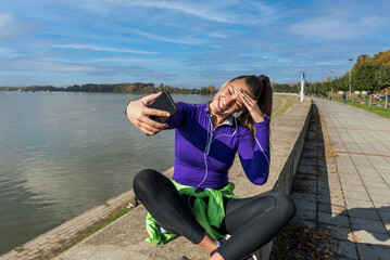 Young sweaty active fitness athlete woman taking a break after outdoor training and taking a selfie with her smartphone for social media to show her success of running and jogging exercises