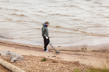 Side view of young man walking along coastline and using metal detector to find coins or valuable relics on sea beach