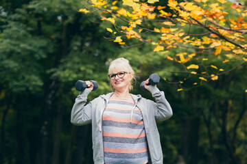 Beautiful senior woman on a walk in the park, performs exercises with dumbbells in her hands
