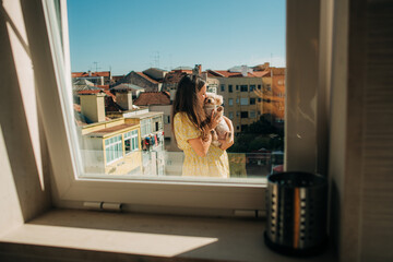 Young brunette pregnant woman holding and hugging a puppy while standing at balcony in Lisbon, Portugal in a sunny summer day. View from a kitchen window