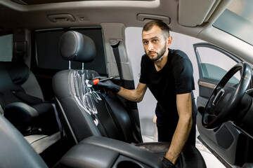 Car detailing and cleaning concept. Young man, car wash worker, wearing protective rubber gloves, cleaning the car seat of the modern vehicle, using special brush and foam, looking at camera
