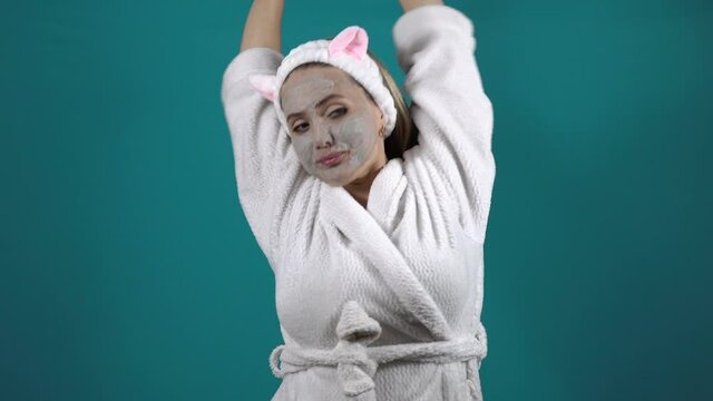 A woman in a white robe, wearing a carbonated bubble clay mask, yawns and stretches