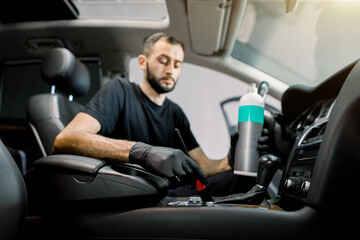 Car interior plastic care, detailing concept. Portrait of young handsome male Caucasian auto service worker, cleaning car interior with soft brush and special professional care milk. Focus on the hand