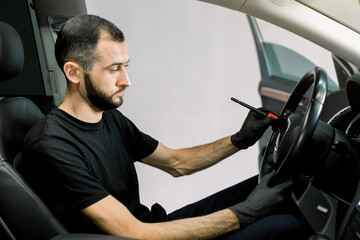 Car detailing and interior care concept. Young man, car wash worker, wearing protective rubber gloves, cleaning the steering wheel of the modern vehicle, using special brush. Selective focus