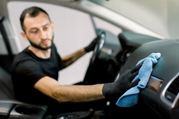 Cleaning service, car detailing concept. Young man, auto service worker, washing a car interior, console and dashboard with microfiber cloth, in a professional car wash. Fpcus on a hand with cloth