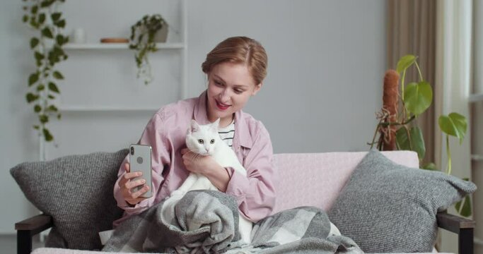 Beautiful young smiling woman blonde girl makes selfie photo with beloved white cat adorable pet for social network uses smartphone mobile phone camera while sitting on sofa in living room at home