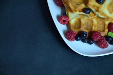 Homemade Belgium Waffles with fresh blueberries for breakfast. White plate, Viennese waffles with raspberries and mint. quick delicious breakfast on black background. top view copy space