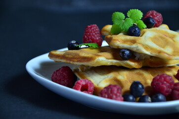 Homemade Belgium Waffles with fresh blueberries for breakfast. White plate, Viennese waffles with raspberries and mint. quick delicious breakfast on black background. top view copy space