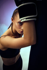 athletic woman in black boxing gloves leaning on punching bag on smoke studio background. woman resting after workout. close-up portrait